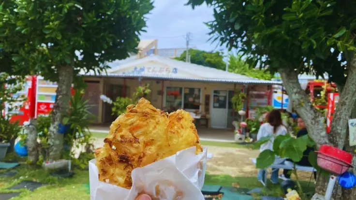 奥武島の大城てんぷら店を背景に揚げたての天ぷらを持つ様子