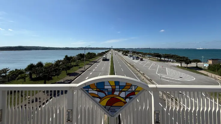海中道路の歩道橋から、まっすぐ伸びる道路と両側の青い海を見渡している様子