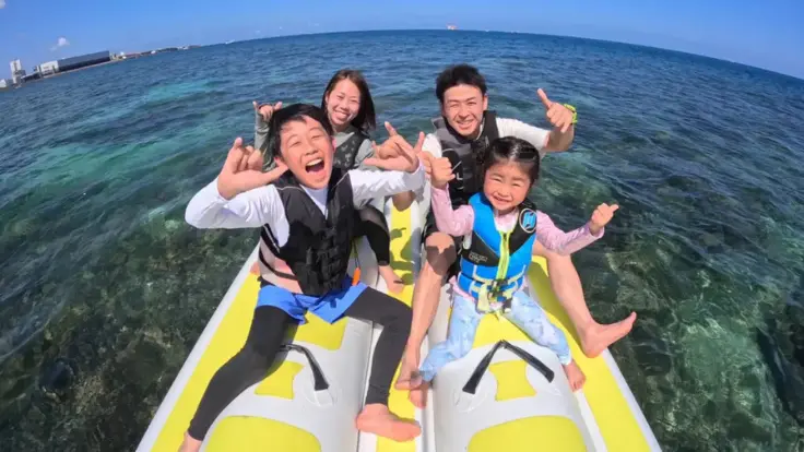 沖縄の海で家族でバナナボートを楽しんでいる様子。みんなで並んで乗り、海の上を走っている体験イメージ。